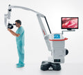 A refurbished Leica M720 / OH5 surgical microscope with a white and gray body, red accents, and an attached monitor displaying a surgical image, with a surgeon in a blue gown operating on the microscope.